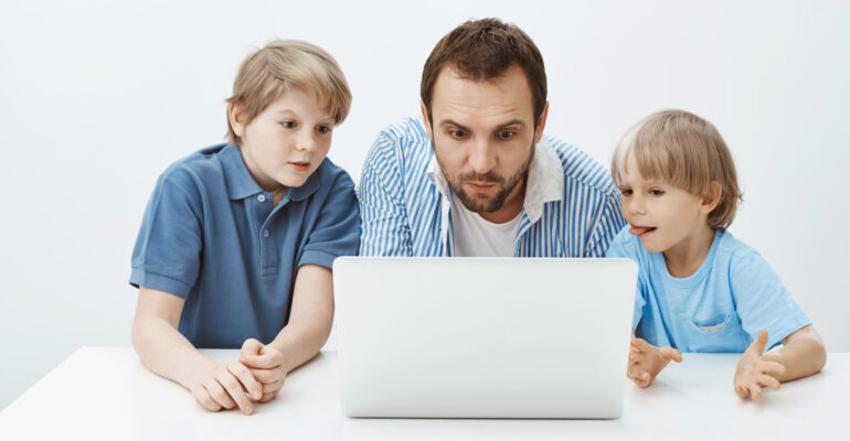 Father watching cartoons with sons. Portrait of enterteined attractive father with young boys sitting at table and looking at laptop screen with interest and curiousity, spending time with family